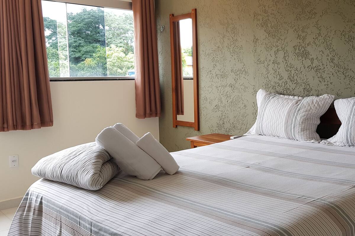A neatly made bed with striped bedding and two pillows, a wooden nightstand, a mirror, and a window with curtains.