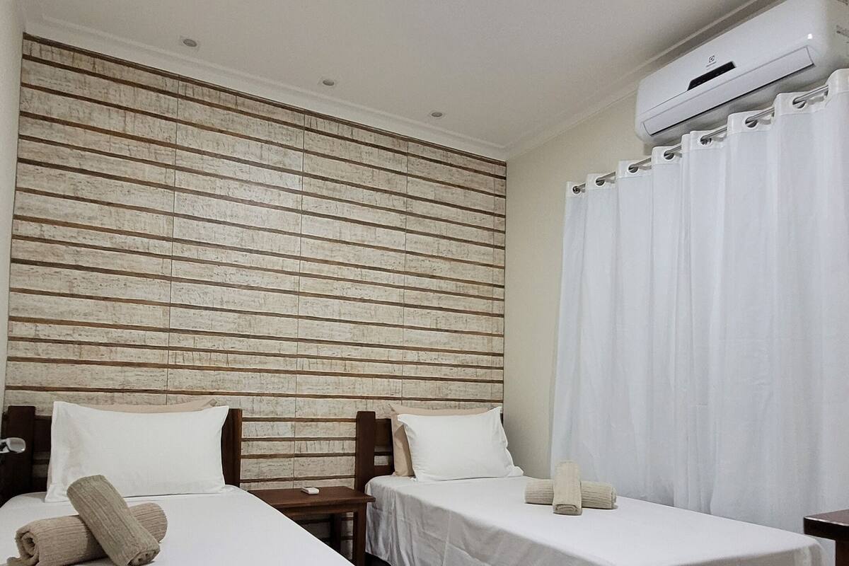 A hotel room with two single beds, a wooden headboard, a white curtain, and an air conditioning unit.