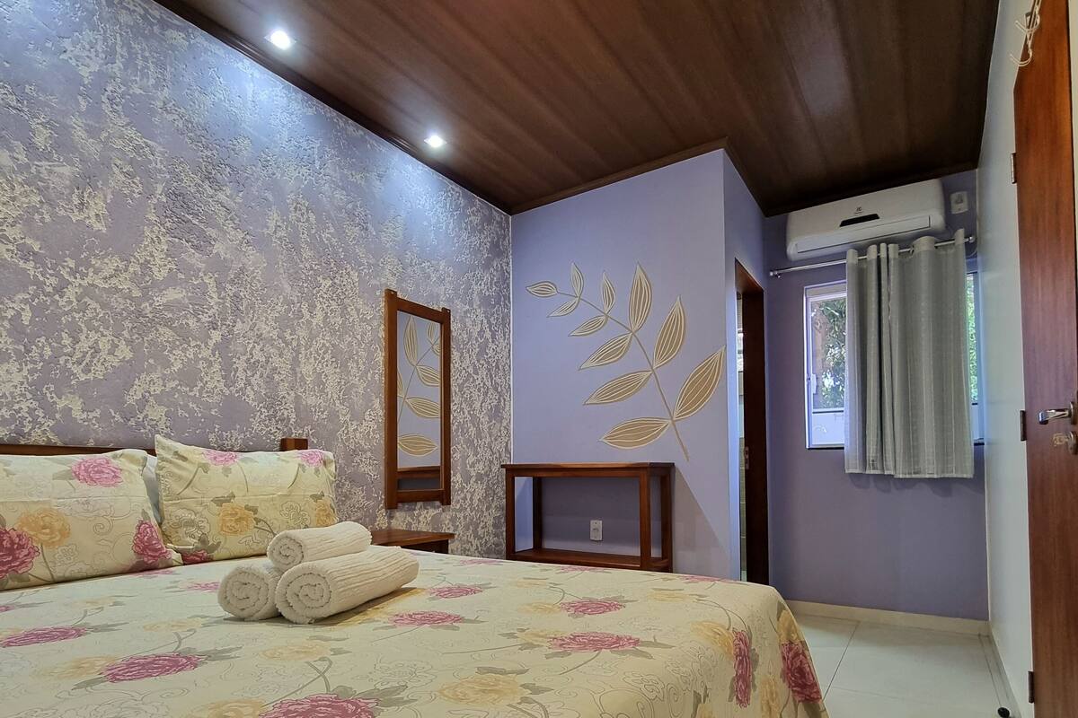 A bedroom with a floral bedspread, a wooden ceiling, a mirror, and a window with curtains.