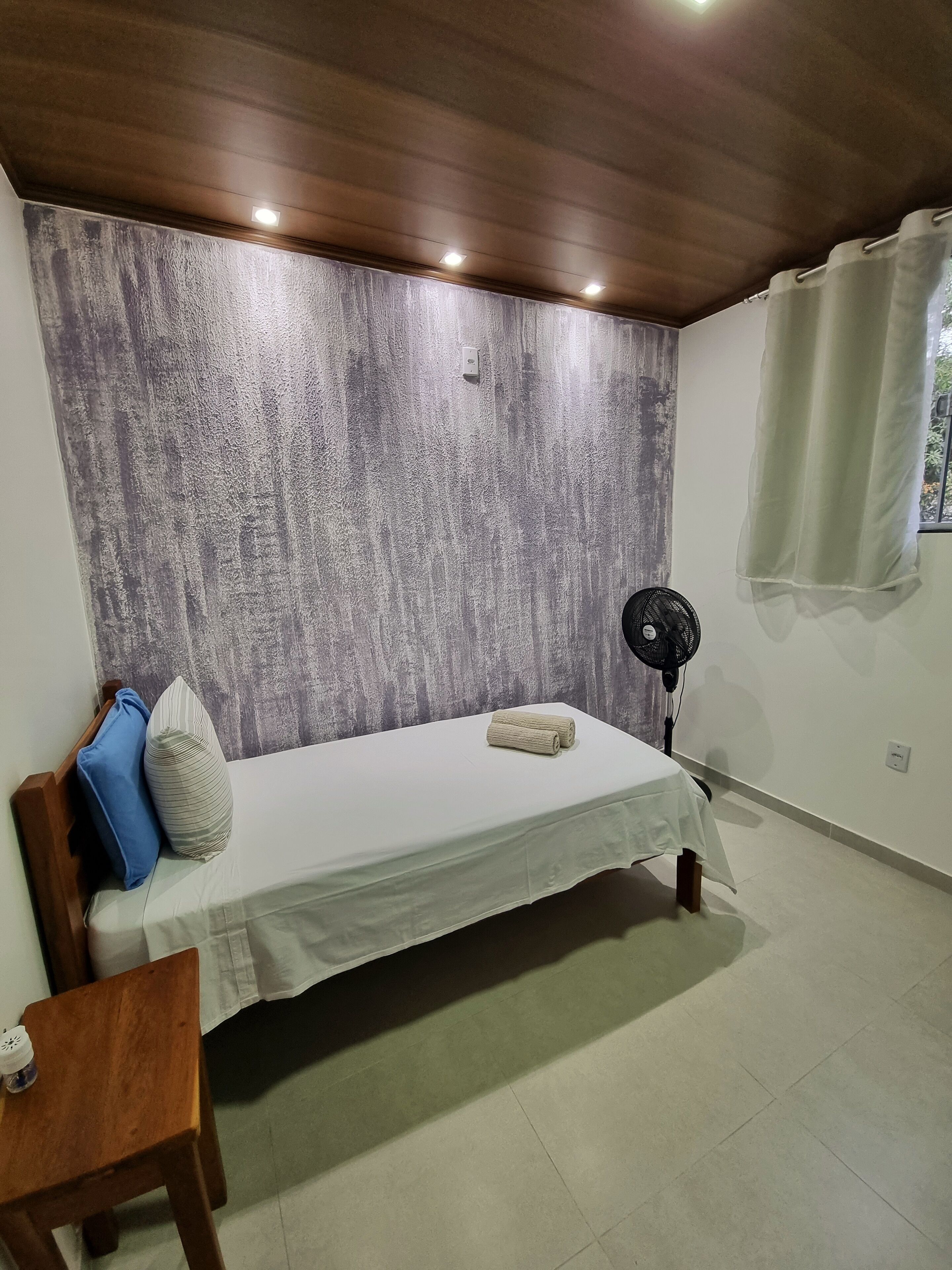 Une chambre à coucher avec un lit, une table de chevet en bois, un ventilateur et une fenêtre avec des rideaux.