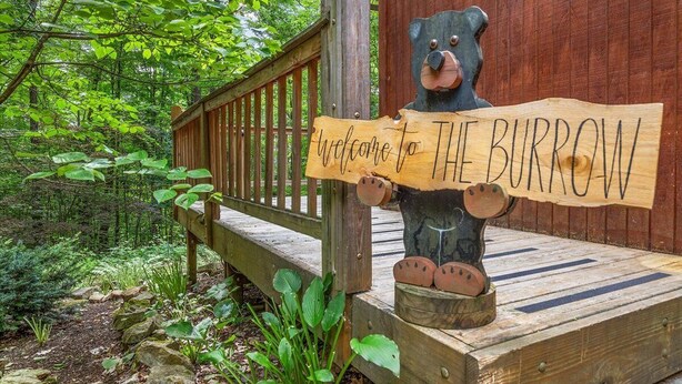 A wooden sign with a bear holding a welcome sign, set on a wooden deck with a forest backdrop.