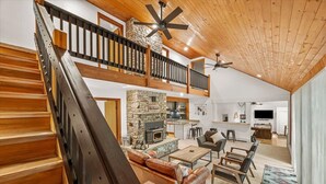 A spacious living area with a stone fireplace, wooden staircase, and a high ceiling with exposed beams.