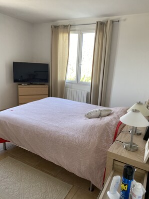 A hotel room with a bed, a TV on a wooden dresser, a window with curtains, a bedside table with a lamp, and a radiator. - Beautiful air-conditioned country villa, sleeps 10, with saltwater pool. (Manosque)