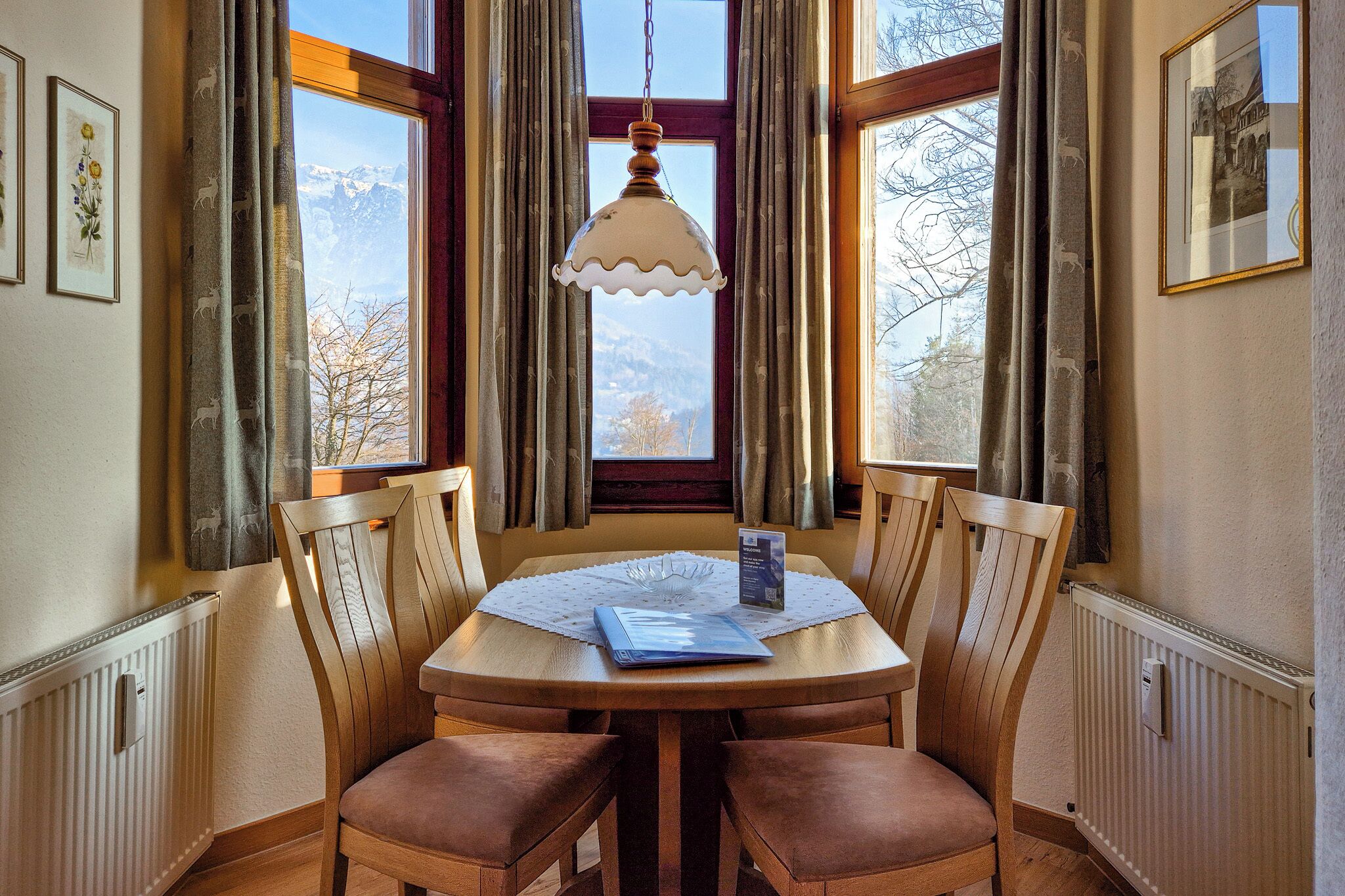 A dining area with a round wooden table, four wooden chairs, a hanging light fixture, and a view of mountains through a window with curtains.