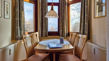 A dining area with a round wooden table, four wooden chairs, a hanging light fixture, and a view of mountains through a window with curtains.