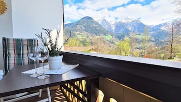 A balcony with a table, two wine glasses, and a potted plant. A mountain view is visible through the window.