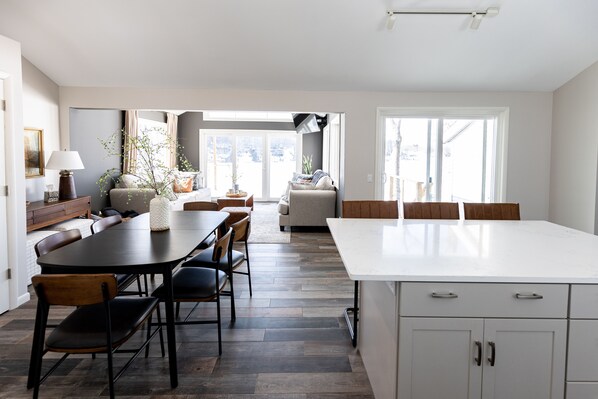 A modern kitchen with a large island, dining area with chairs, and a view of the city through large windows. - Allsports lakehouse w/private beach/dock, firepit, grill, sunset view! (Belding)