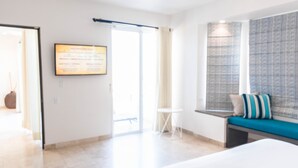 Hotellihuone, jossa on sänky, pieni pöytä, tyynyillä varustettu penkki, ikkuna, jossa on verhot, ja ovi, joka johtaa toiseen huoneeseen.