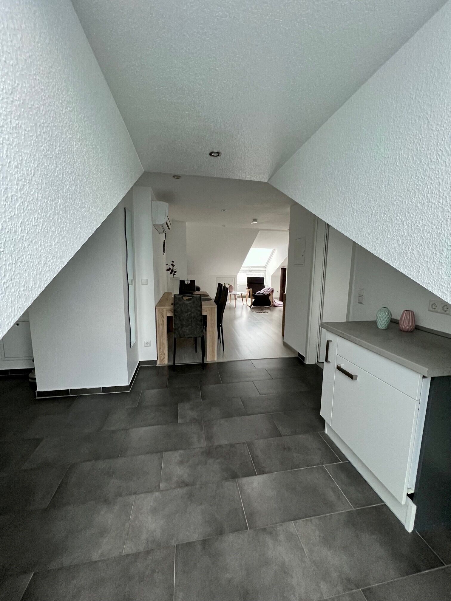 A spacious attic room with a sloped ceiling, a dining area with a table and chairs, and a kitchenette with cabinets and a countertop.