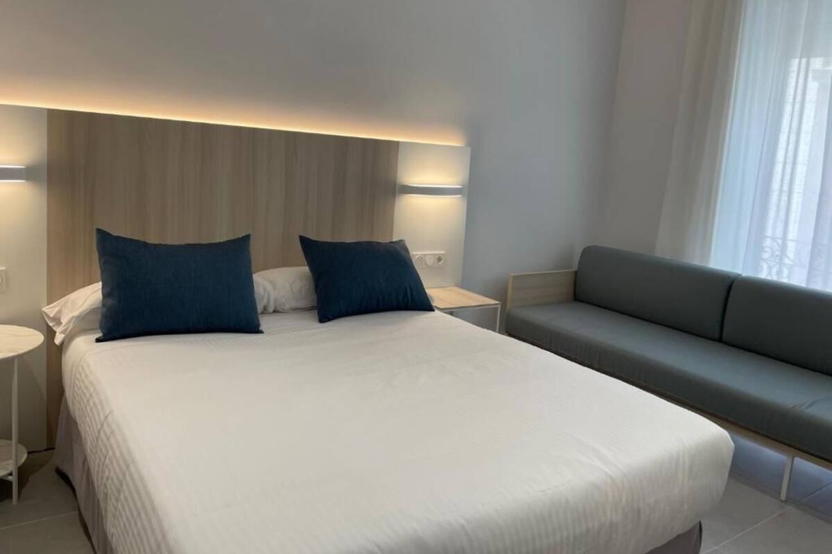 Ein modernes Hotelzimmer mit einem großen Bett, zwei blauen Kissen, einem grauen Sofa und einem Nachttisch.