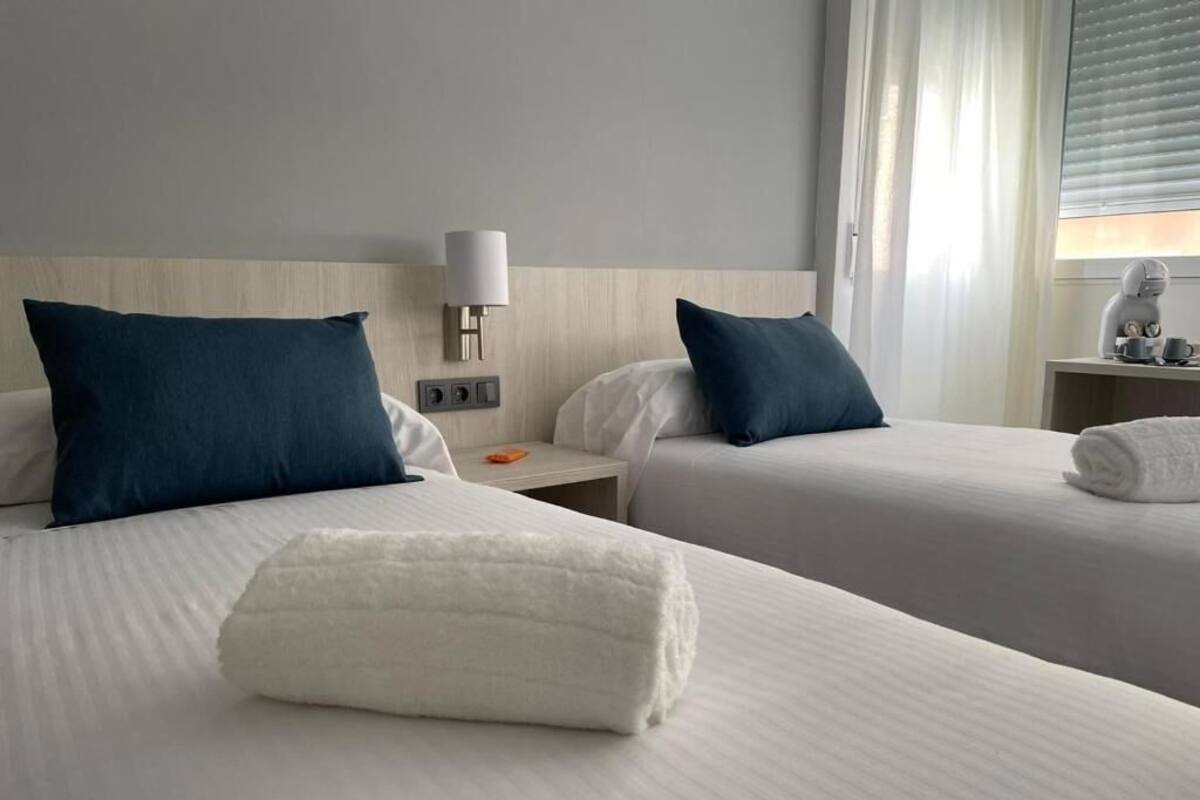 Ein Hotelzimmer mit zwei Betten, jedes mit einer weißen Tagesdecke und einem dunkelblauen Kissen. Ein Nachttisch mit einer Lampe und ein kleiner Tisch mit einer Kaffeemaschine.