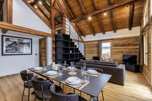 A dining area with a long table set for four, a staircase, a living room with a sofa and TV, and wooden walls.