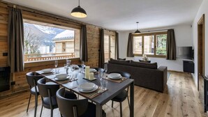 Un comedor con una mesa dispuesta para cuatro personas, un sofá y un televisor en una habitación con paredes de madera y ventanas amplias.