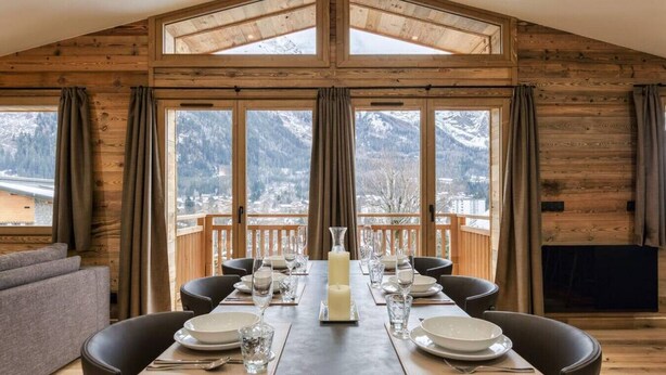 Un comedor con una mesa larga dispuesta para seis comensales, ventanas amplias con cortinas y vistas a montañas nevadas.