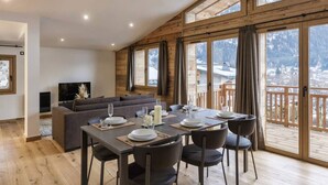 Un comedor con una mesa dispuesta para cuatro personas, un sofá y un televisor en una habitación con paredes de madera y ventanas amplias.