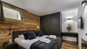 A modern bedroom with a wooden wall panel, a bed with dark bedding, a bedside table with a lamp, and a small bedside table with a candle.