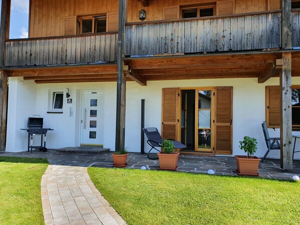 A two-story wooden building with a balcony, a door with a number, a grill outside, and potted plants.