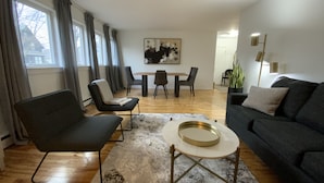 A modern living room with a dark grey sofa, a round coffee table, and a dining area with a wooden table and chairs. - Beautiful Brighton/Charlottetown; 1300 sq ft. apartment with AC and large deck.  (Charlottetown)