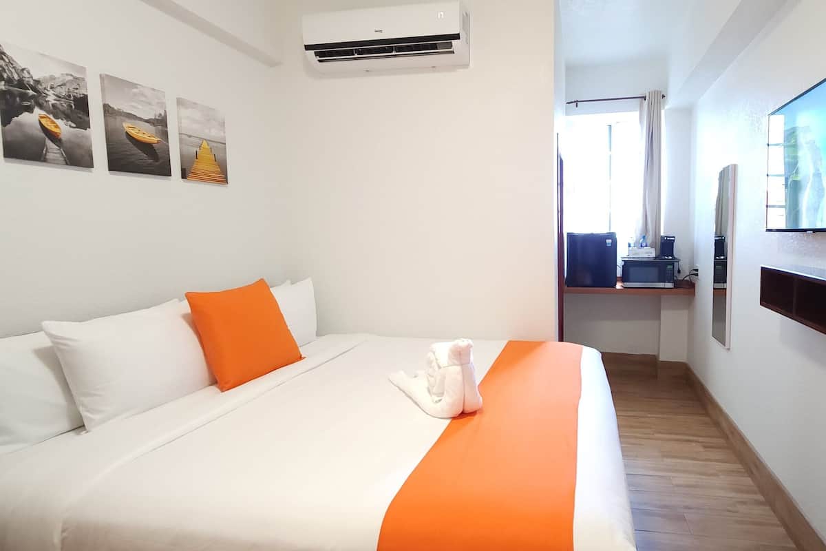 A hotel room with a bed, a flat-screen TV, a wall-mounted air conditioner, and a window with curtains.