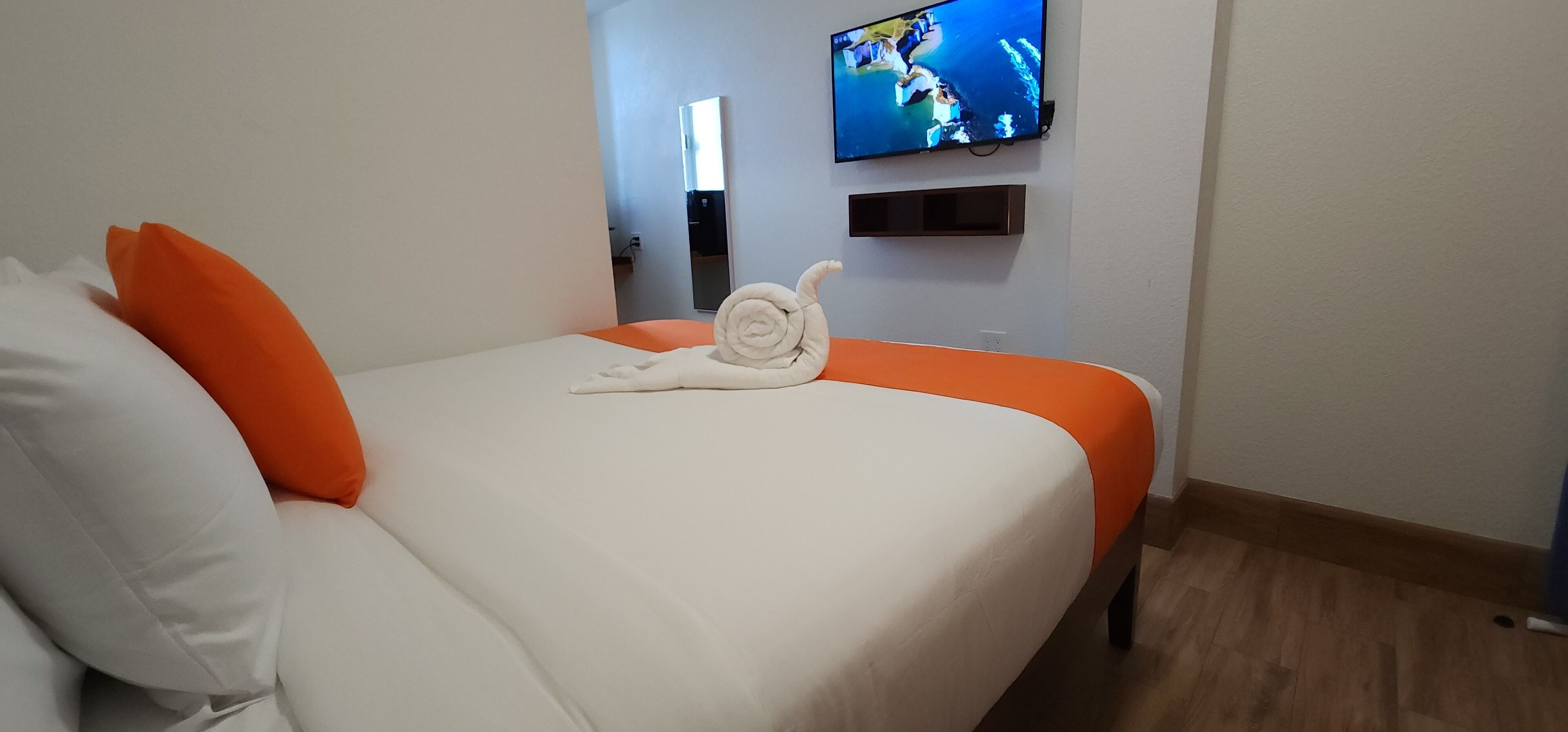 Una habitación de hotel con cama, ropa de cama naranja, almohada blanca, toalla enrollada, televisor en la pared y suelo de madera.