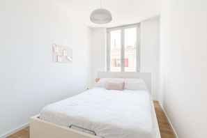 A minimalist bedroom with a single bed, a window with a view of buildings, and a wall-mounted light fixture. - MaisonMars Le Cosy Blancarde (Marseille)
