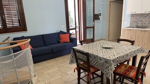 A living room with a sofa, a dining table with chairs, a small kitchen area, and a window with blinds. - Villa with pool and with sea view (San Vito Lo Capo)