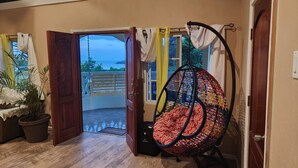 A room with a wooden floor, a hanging chair, a potted plant, and a view of the sea through an open door.