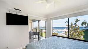 Ein Schlafzimmer mit einem großen Bett, einem flachbildfernseher an der Wand und einem Balkon mit Blick auf das Meer und Palmen.