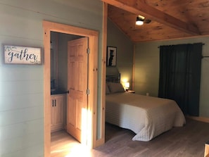 Een slaapkamer met een bed, een nachtkastje, een wandlamp, een deur naar een badkamer, en een bordje met de tekst 'gather' aan de muur.