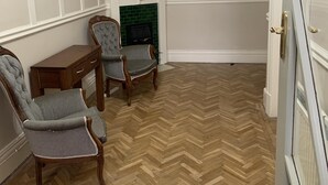 A room with a herringbone wooden floor, a fireplace with a green tiled surround, two upholstered chairs, a wooden side table, and a mirror above the fireplace.