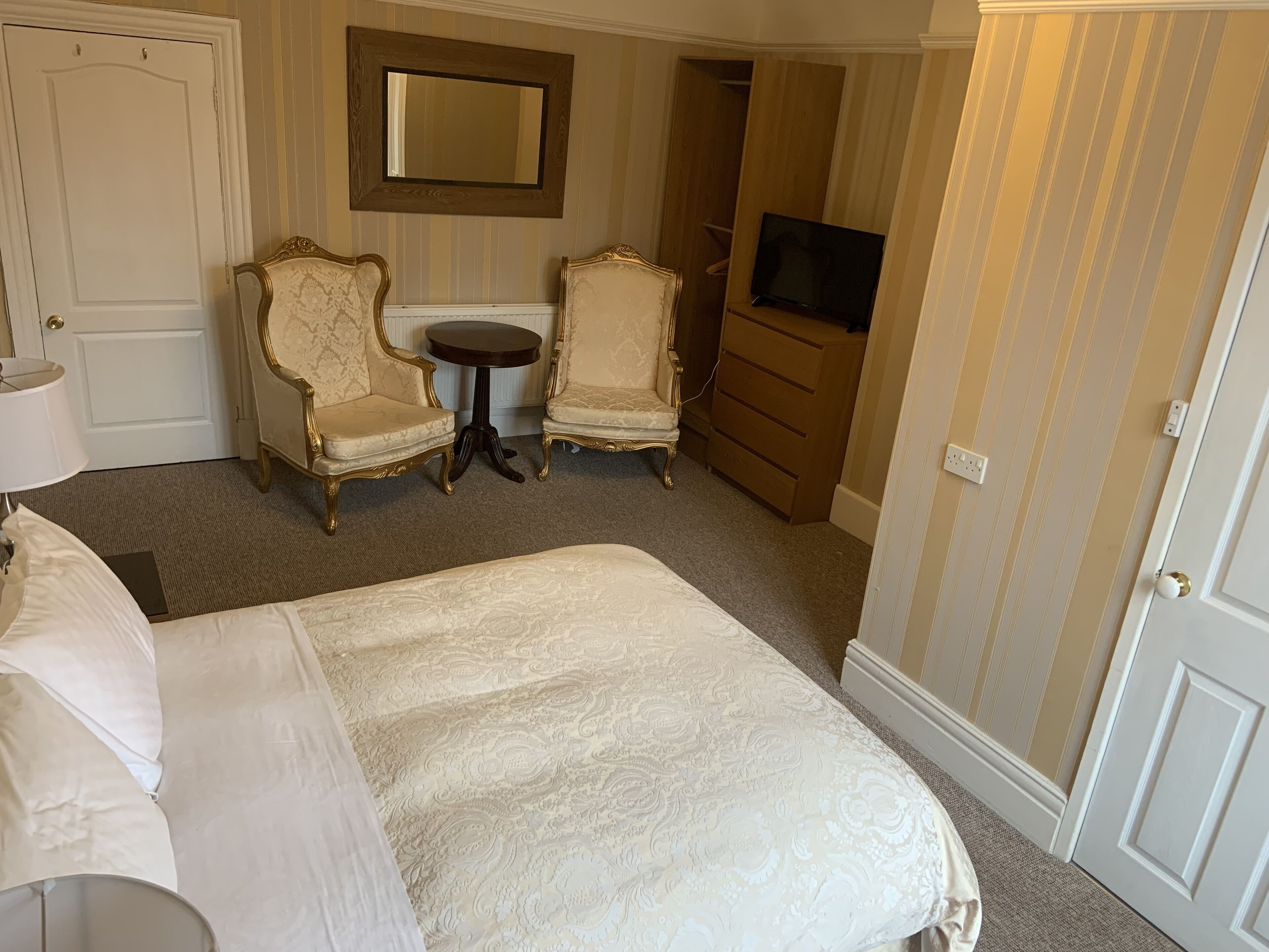 A hotel room with a bed, two ornate chairs, a small round table, a dresser, a television, and a mirror on the wall.