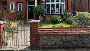 A two-story brick house with a bay window, a front door, and a small garden with flowers and shrubs.
