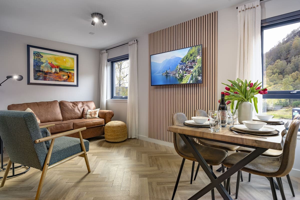 Un salon moderne avec un canapé en cuir marron, un fauteuil bleu et une table à manger en bois, mise avec des assiettes, des verres et un vase de tulipes rouges.
