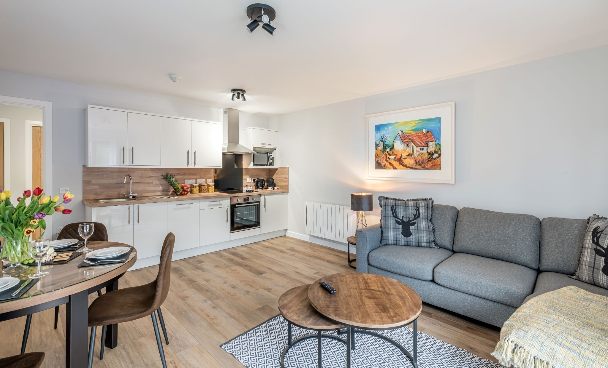 A modern kitchen with white cabinets, a wooden countertop, and a built-in oven. There is a dining area with a table set for two, a grey sofa, and a painting on the wall.