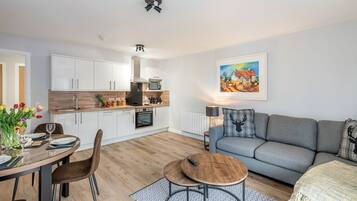 A modern kitchen with white cabinets, a wooden countertop, and a built-in oven. There is a dining area with a table set for two, a grey sofa, and a painting on the wall.