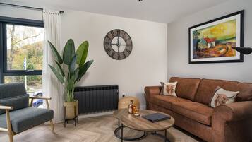 A living room with a grey armchair, a brown sofa, a wooden coffee table, a large potted plant, a wall clock, and a framed painting.