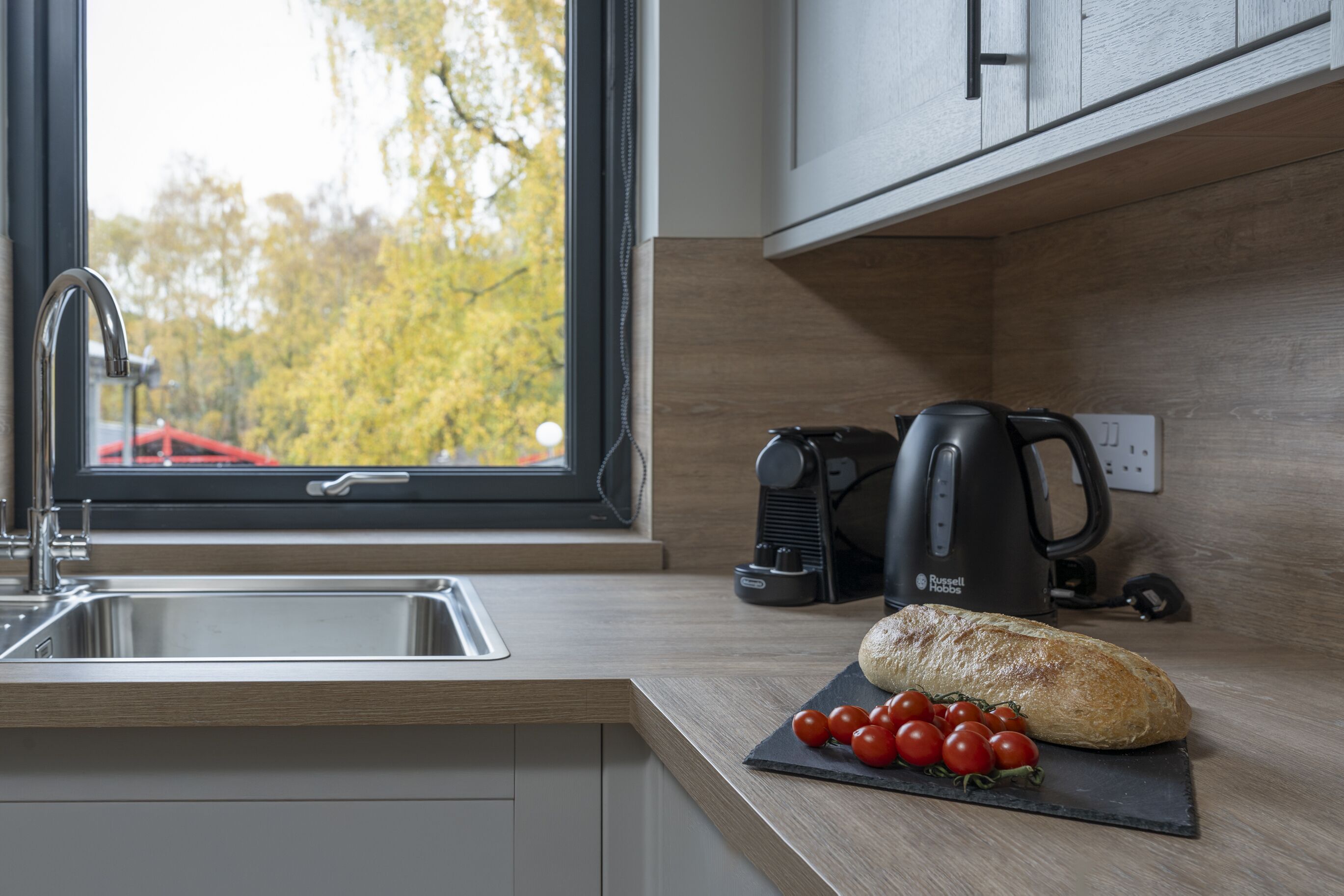 A kitchen with a modern design, featuring a stainless steel sink, a kettle, and a loaf of bread on a cutting board with tomatoes.