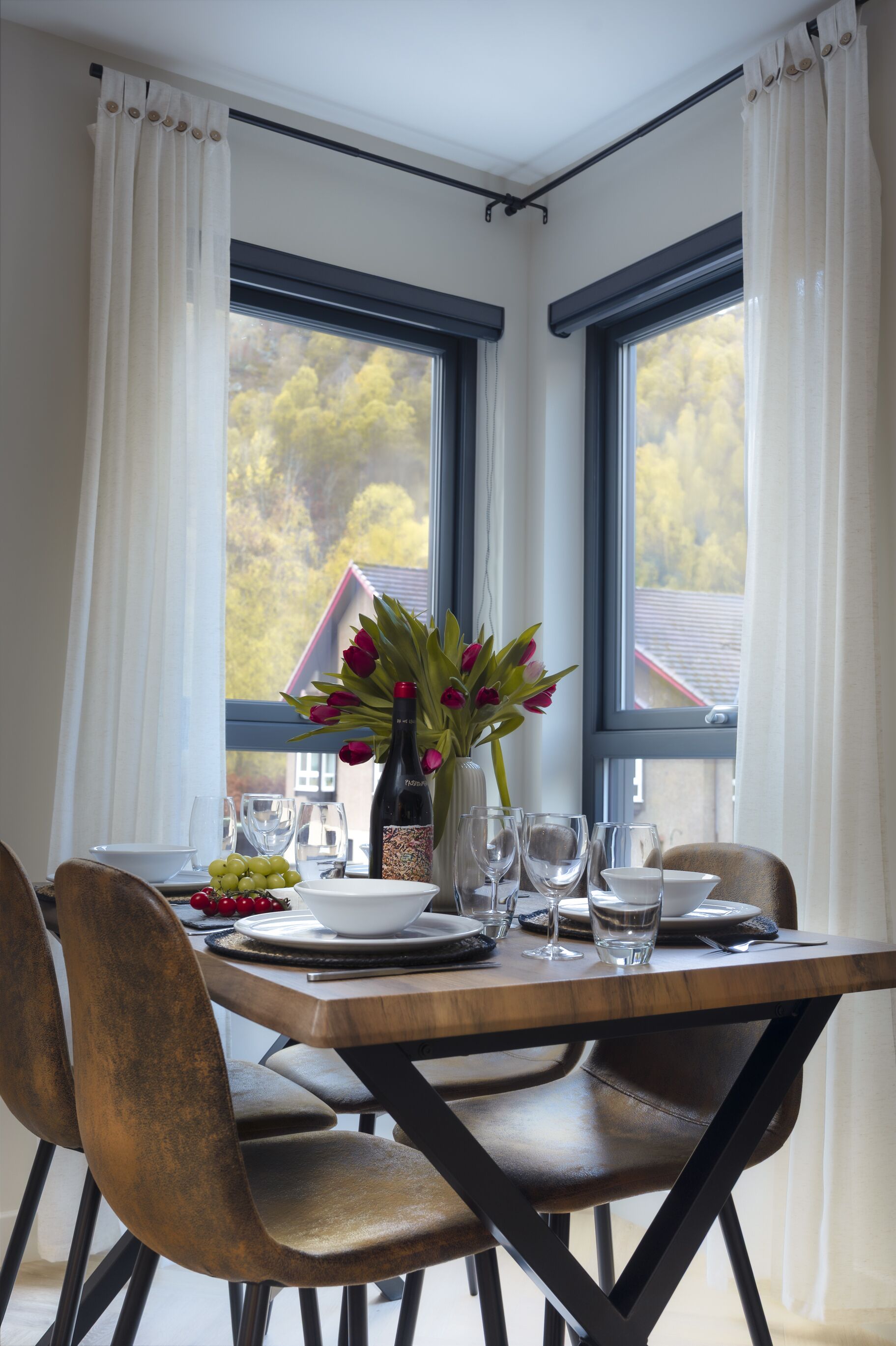 A dining table set with plates, glasses, and a bottle of wine, positioned near a window with curtains.