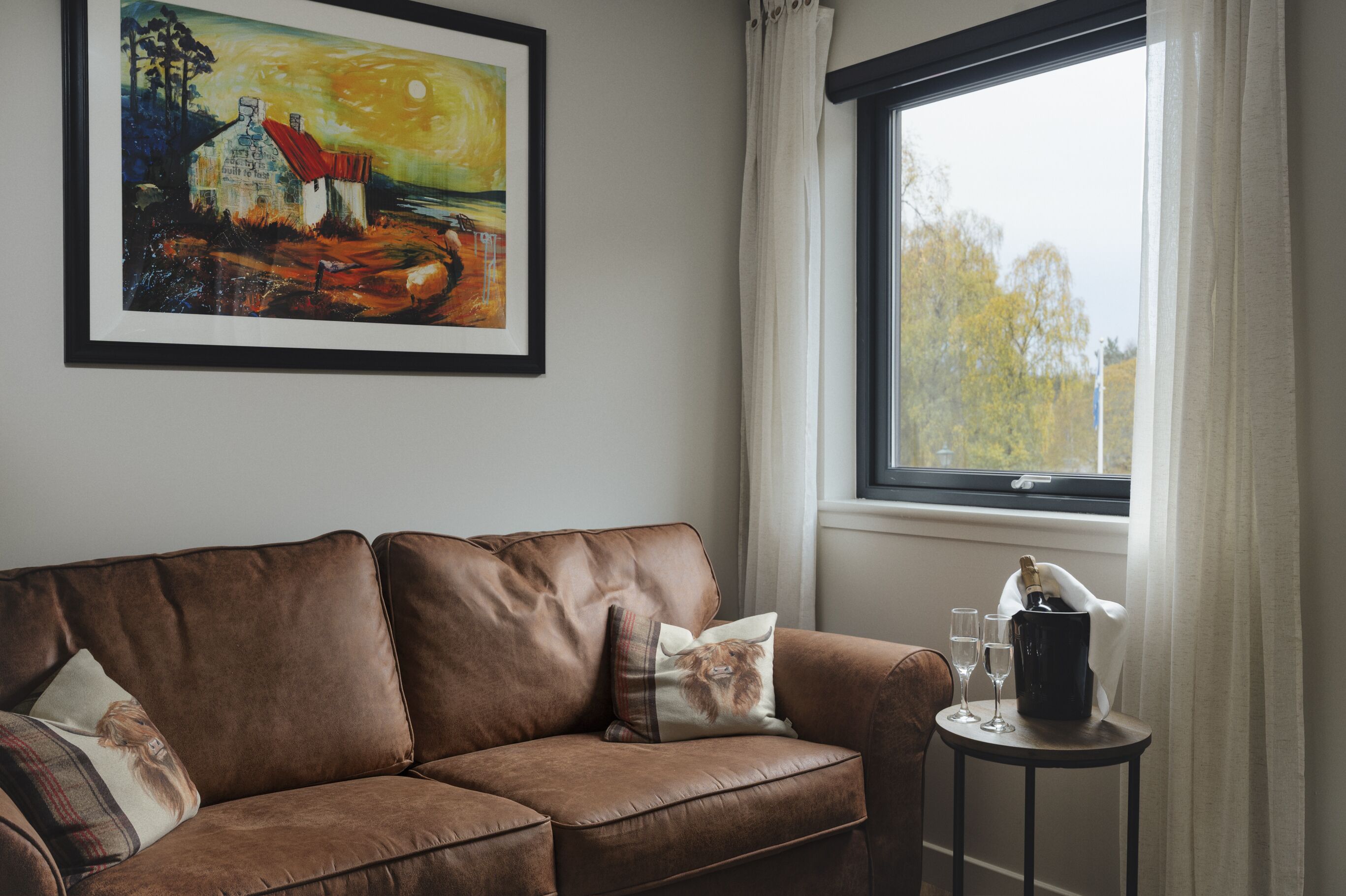 A brown leather sofa with two pillows, a framed painting above it, a window with curtains, and a small table with a bottle and glasses.