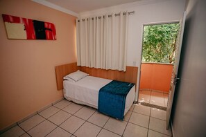 A small, single-bed room with a wooden headboard, a white bedspread, and a blue blanket. There is a window with white curtains, a door leading to a balcony, and a red and black abstract painting on the wall. - Diroma Fiori Hotel - BVTUR (Caldas Novas)