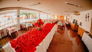 A spacious banquet hall with a long table covered in red cloths, large windows, and a kitchen area visible to the right. - Diroma Fiori Hotel - BVTUR (Caldas Novas)