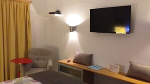 Hotellihuone, jossa on sänky, kirjoituspöytä lampun kanssa, tuoli, seinälle asennettu televisio ja verho.