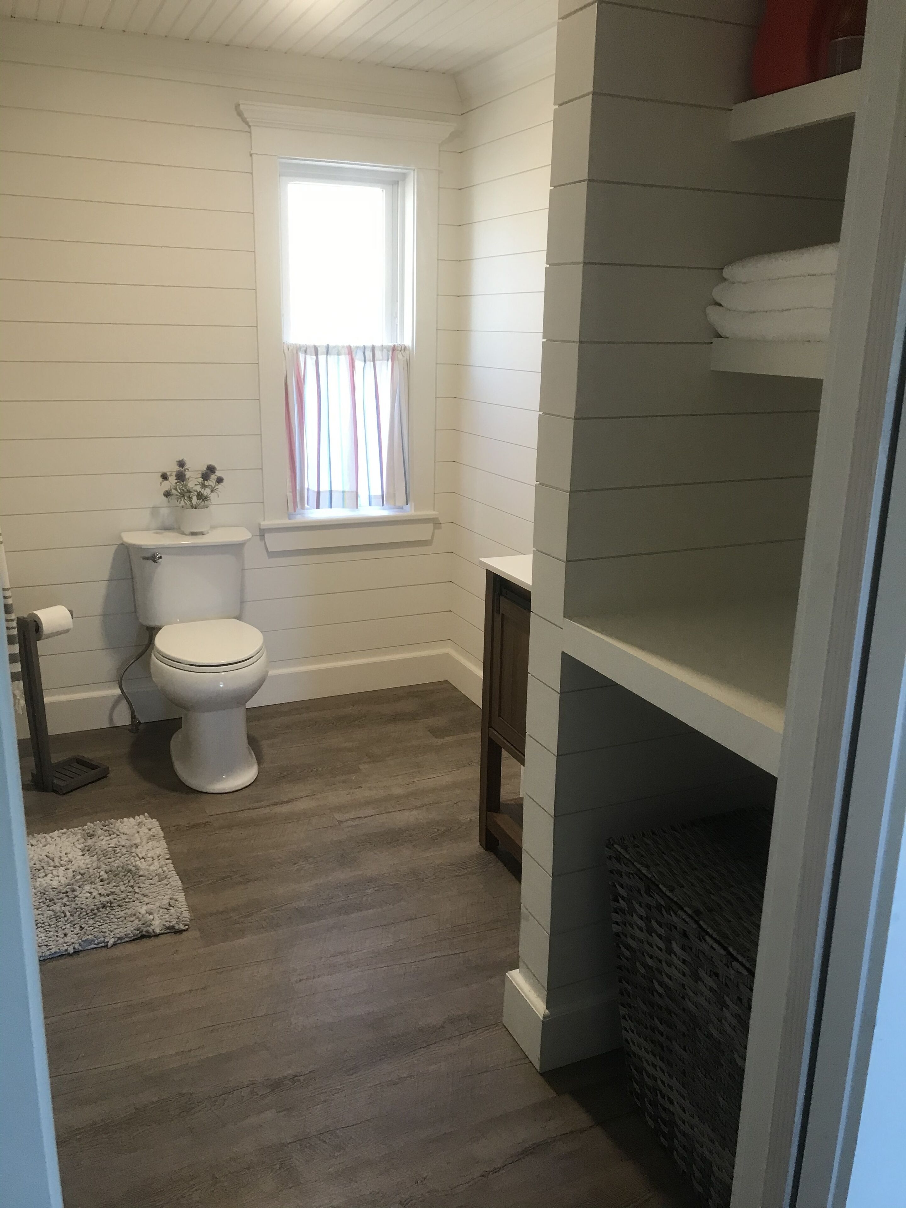 Ein Badezimmer mit Toilette, einem fensterbelaubten Fenster, Holzboden und einer integrierten Ablage mit Handtüchern.