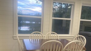 Een eetgedeelte met een houten tafel en witte stoelen, grote ramen met uitzicht op een meer, en een blauwe bank op de achtergrond.