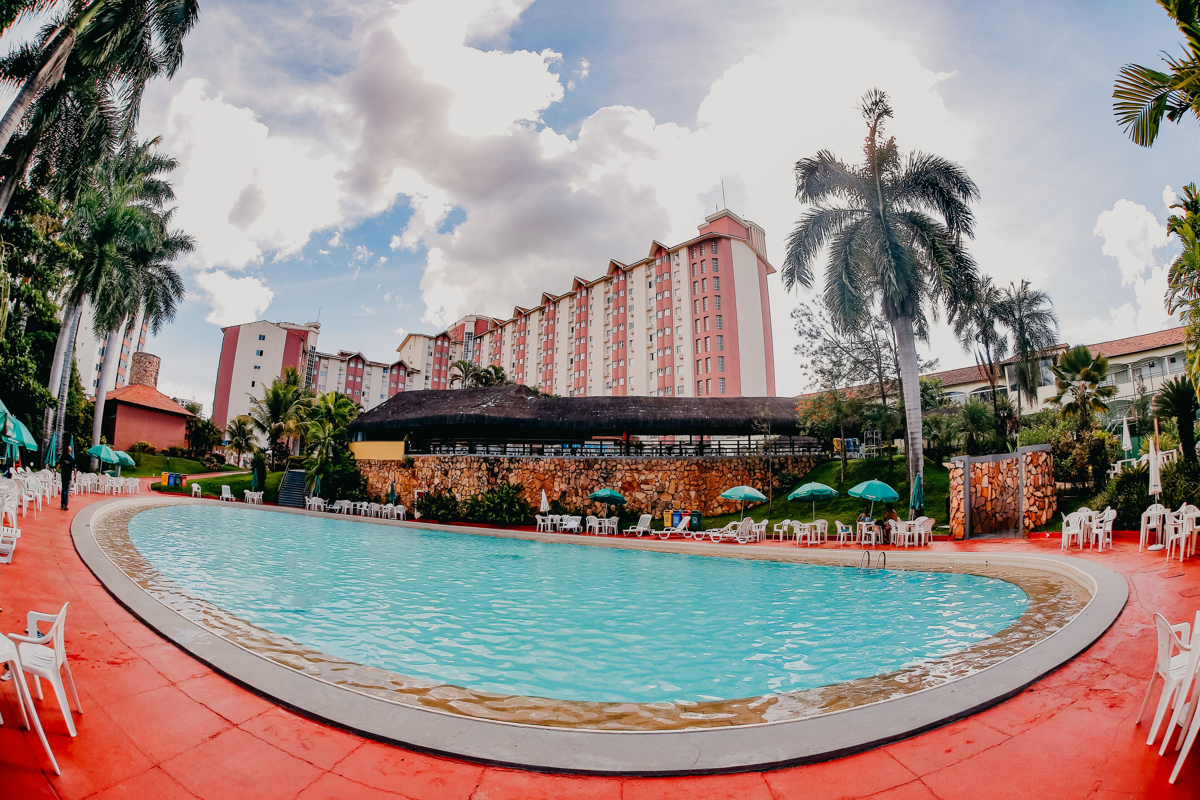 Una piscina con sillas de descanso y sombrillas, rodeada de palmeras y un edificio de hotel.