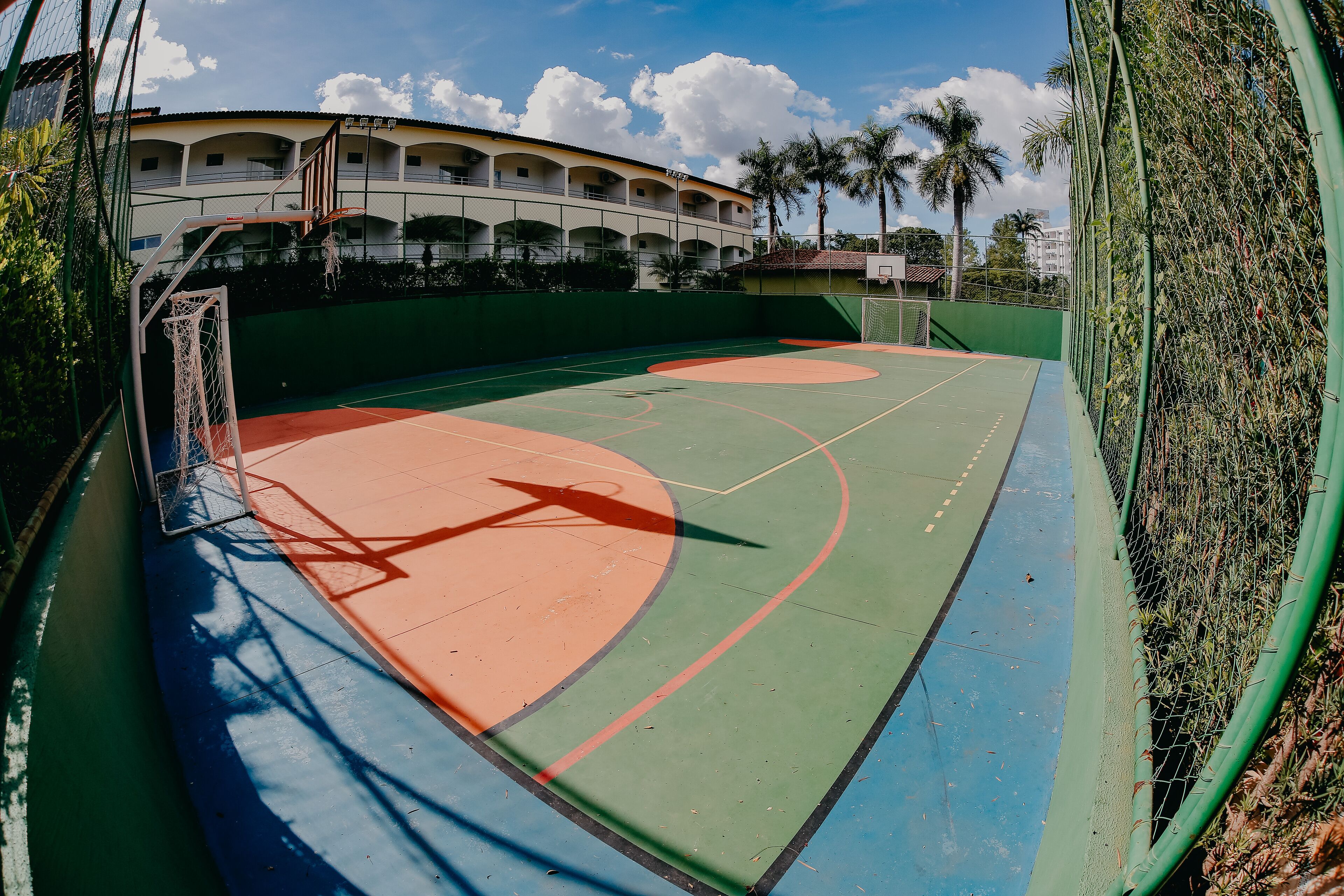 An outdoor basketball court with a green and orange playing surface, surrounded by a green fence and palm trees.