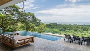 Zona de piscina con una silla colgante y dos sillas de metal, con vistas a un paisaje verde exuberante y el océano.