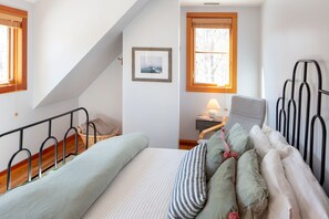 A bedroom with a bed, a chair, a nightstand, a lamp, a picture on the wall, and a window with a view of trees.