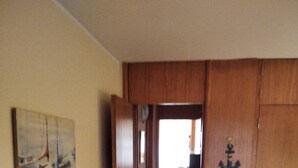 A room with a wooden door leading to a hallway, a painting of sailboats on the wall, and a bed with a red blanket.