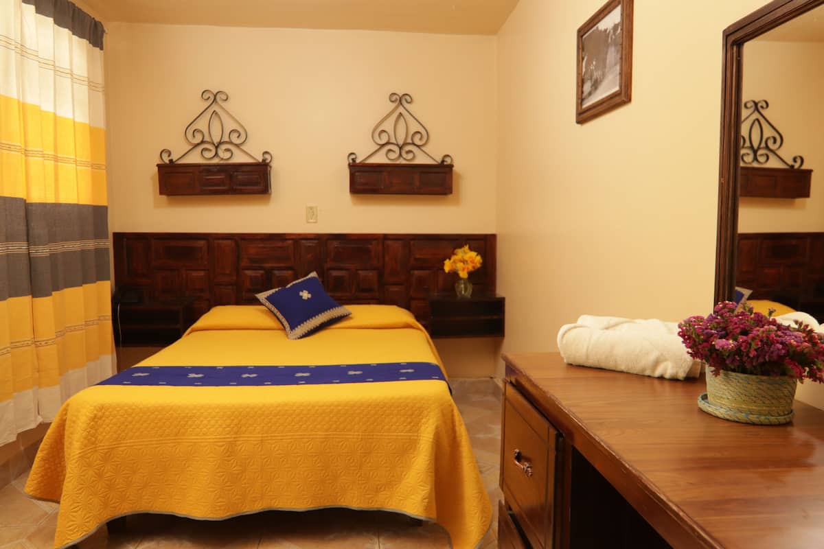 Ein Hotelzimmer mit einem Bett, einem Holzkopfteil, einem Nachttisch mit Blumenvase, einem Spiegel und einem an der Wand befestigten Regal.